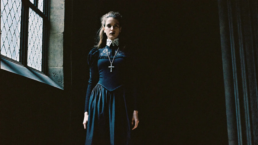Woman in a long black Victorian-style dress with a high lace collar and silver cross pendant in the light of a gothic cathedral window